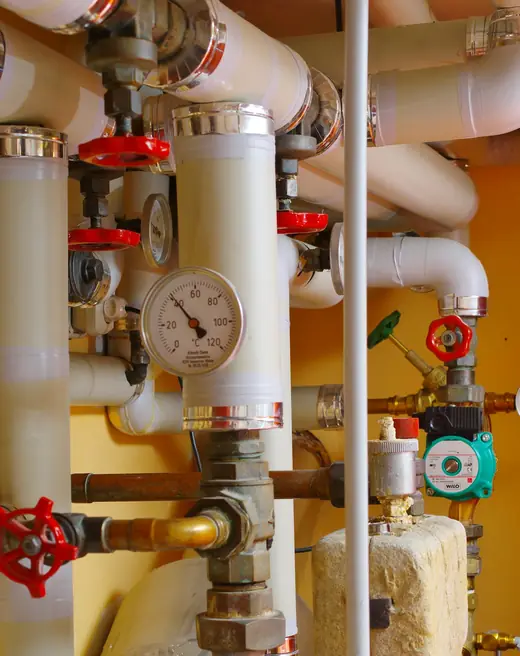 Insulated heating pipes with red handwheel valves and a round pressure gauge, fixed to a yellow wall.