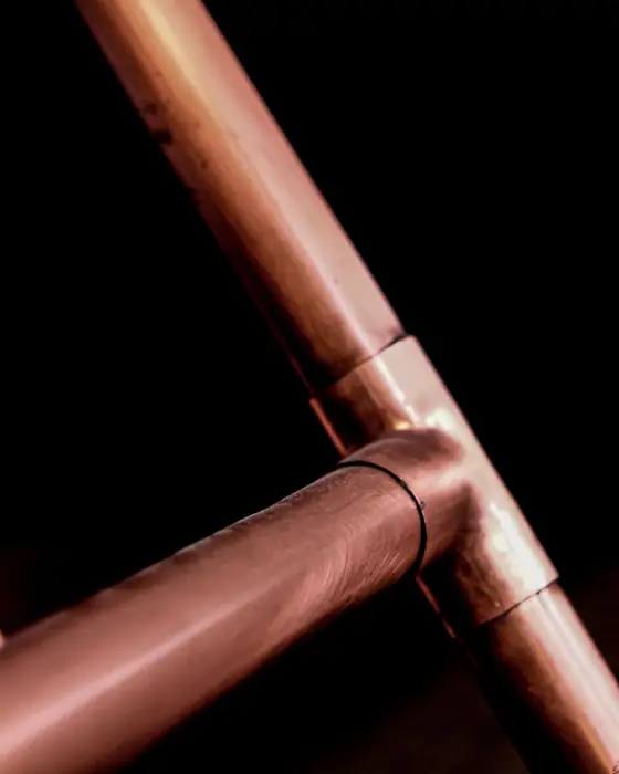 A copper tee joint where two lengths of copper pipe meet, lit against a black ground.